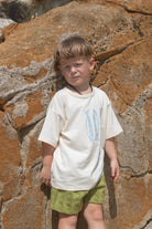 Child wearing a white t-shirt with blue fish and green shorts standing against a rocky wall.