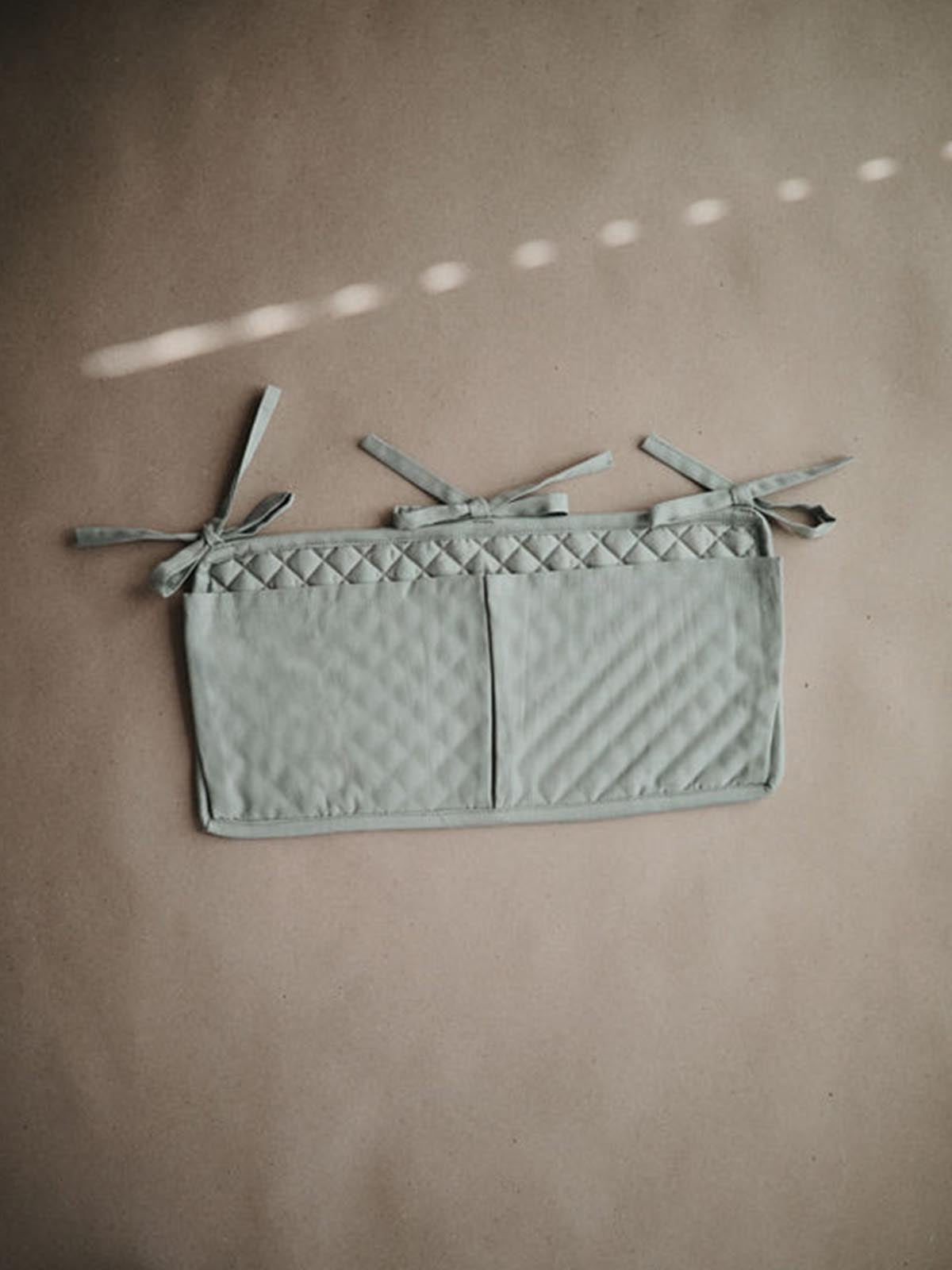 Mushie Roman Green crib pocket hanging on a crib, made of soft fabric, designed for storing small baby essentials.